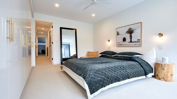 Bright, contemporary bedroom with a large upholstered bed dressed in dark textured bedding, pale carpet, white walls, built-in wardrobes with brass handles, bedside stump table, framed artwork, wall sconces, and a full-height mirror. A hallway and adjoining spaces are visible through the open doorway.