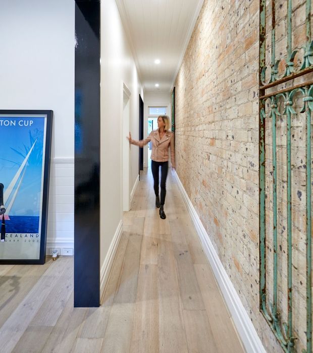 A narrow residential hallway with pale timber flooring, white walls and ceiling, recessed downlights, and a prominent exposed brick feature wall. A dark vertical structural or joinery element separates the corridor from an adjacent room, where framed artwork is visible. Decorative aged metal screening appears along the right edge, and a person is walking through the hall.