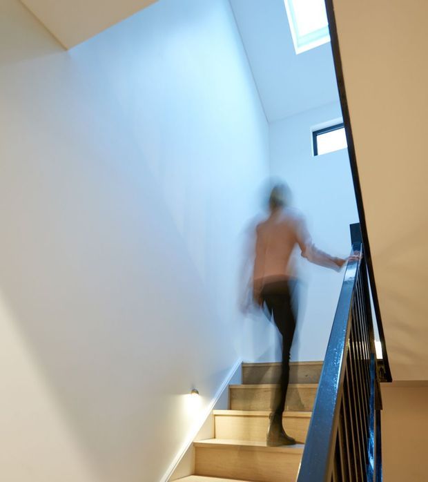 Interior view looking up a modern stairway with light timber treads, white walls, dark metal balustrade, and skylights bringing in natural light. A motion-blurred person is ascending the stairs.