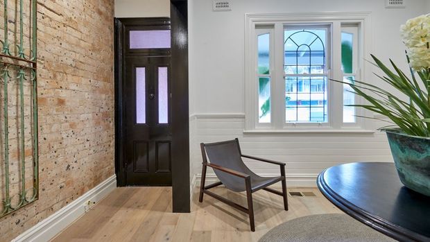 A refined entryway or sitting area with exposed light brick walls, pale timber flooring, white decorative wall panelling and trim, a dark timber front door, and a leadlight-style window. A leather sling armchair and round dark table with a large planter create a small seating vignette.