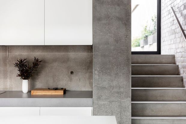Minimalist contemporary interior showing a concrete stairway beside a built-in low console and cabinetry. The space features a raw grey concrete structural column and stair surfaces, white flat-panel joinery, a grey concrete-look backsplash and benchtop, and exposed white brickwork near the stair landing.