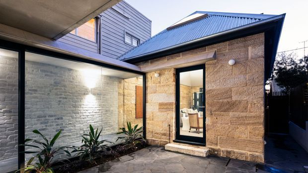 A compact enclosed courtyard beside a single-storey stone-clad building, with a dark pitched metal roof, black-framed glazing and a paved floor. A planted garden bed runs along a pale brick wall, while a bathroom or interior space is visible through the doorway.