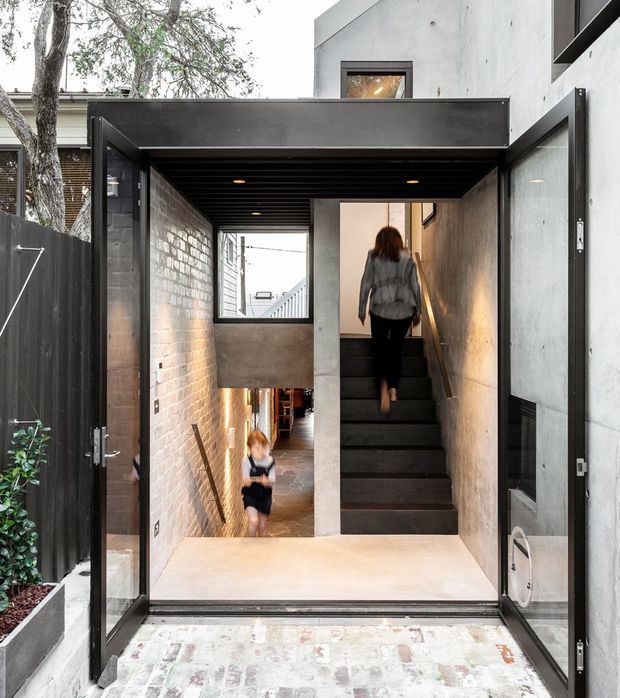 A contemporary residential entry viewed from outside through open black-framed doors. The compact passage features exposed brick walls, raw concrete surfaces, a dark slatted ceiling, and a central stairway leading upward and downward. Warm recessed lighting and glimpses of people add scale, while a small planted area sits beside the entrance.