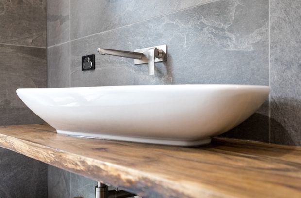 Close-up of a contemporary bathroom vanity featuring a white oval vessel basin mounted on a thick rustic timber benchtop. The wall behind is clad in large grey stone-look tiles, with a wall-mounted chrome tap and a black electrical outlet visible.