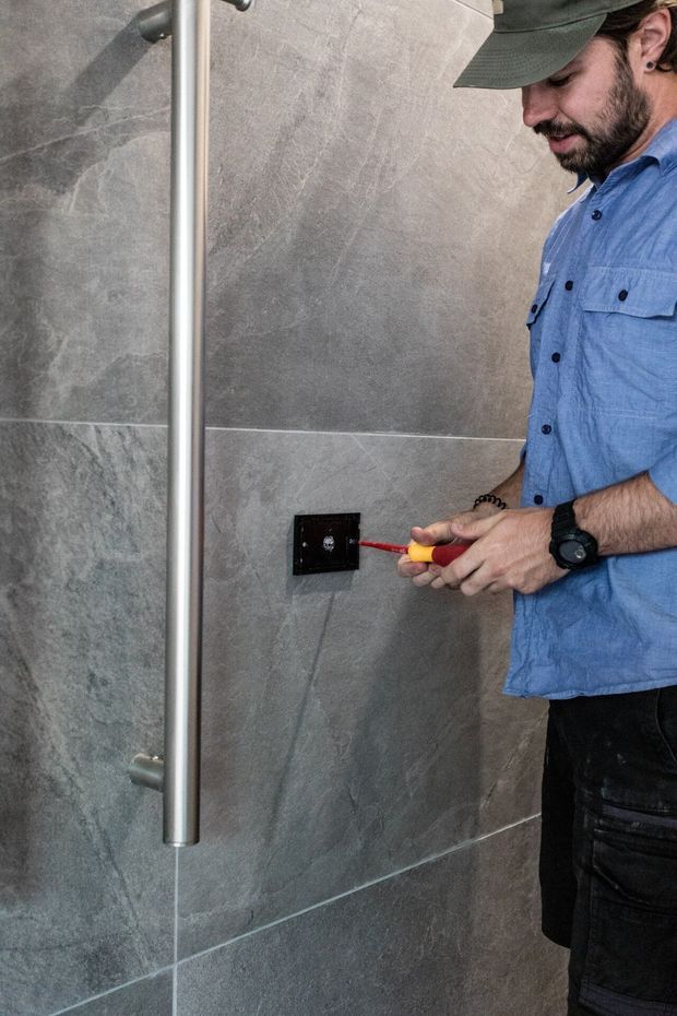 A person using a screwdriver to work on a black electrical switch or outlet mounted on a grey stone-look tiled wall. A vertical metal shower rail or grab bar is visible at the left, indicating a bathroom or wet-room setting. The image focuses on installation or electrical maintenance rather than room styling.