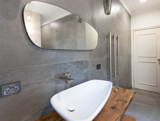 A refined bathroom featuring a freestanding white bathtub set into a thick rustic timber platform, large-format grey stone-look wall and floor tiles, a slim wall-mounted basin tap, an organically shaped black-framed mirror, a vertical heated towel rail, and a white panelled door. The space combines contemporary fixtures with traditional detailing and warm natural wood.