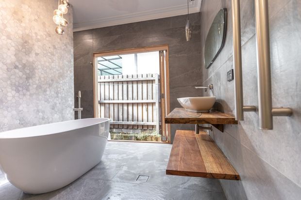 Contemporary bathroom featuring a freestanding white bathtub, vessel basin on a thick timber vanity shelf, large-format grey wall and floor finishes, a textured mosaic feature wall, frameless mirror, and warm pendant lighting. Timber-framed glazing opens toward a fenced exterior area.