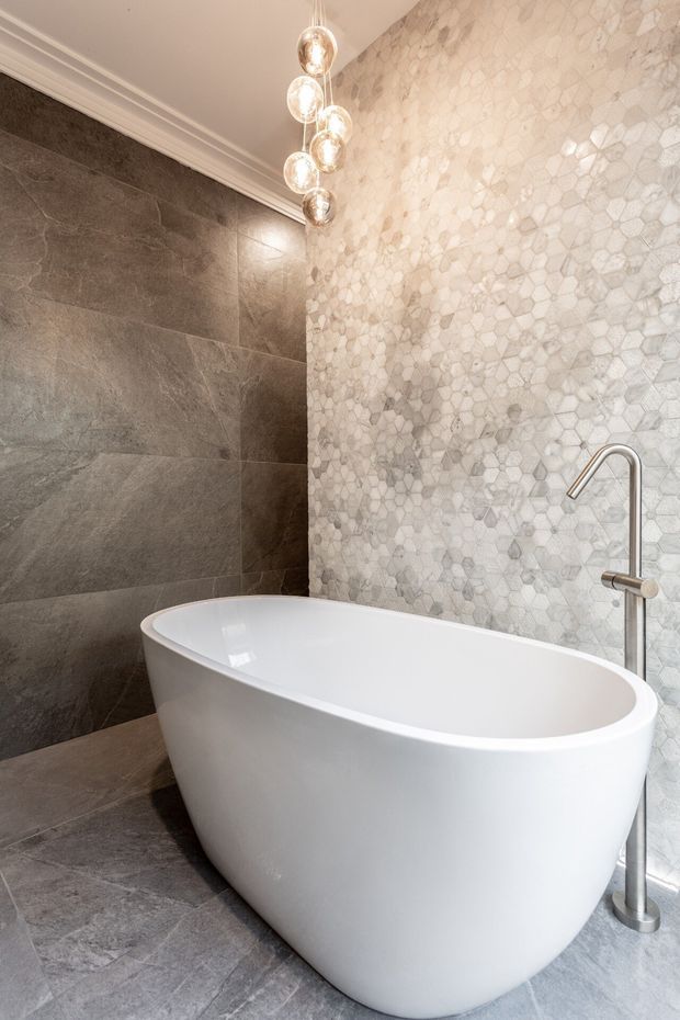 A luxurious contemporary bathroom featuring a freestanding white oval bathtub, a tall brushed-metal floor-mounted tap, large dark grey stone-look wall and floor tiles, and a pale hexagonal mosaic feature wall. A cluster of spherical pendant lights hangs above the bath, with decorative ceiling moulding adding a refined touch.
