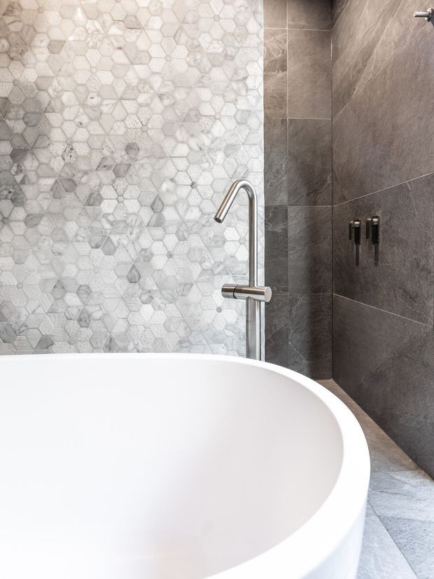 Close-up view of a contemporary bathroom featuring a freestanding white oval bathtub, chrome floor-mounted bath filler, and dark grey large-format wall tiles. A light grey and white hexagonal mosaic tile feature wall contrasts with the darker tiled shower wall and matching floor.