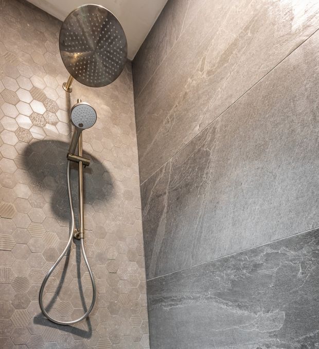 Close-up view of a contemporary walk-in shower with a large round overhead rain shower and handheld shower on a vertical rail. The shower walls combine textured pale hexagonal mosaic tiles with large grey stone-look wall tiles, complemented by brushed metallic fixtures.