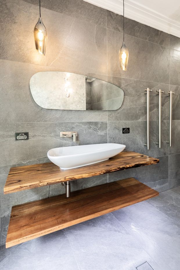 A refined contemporary bathroom featuring large grey stone-look wall and floor tiles, a freestanding white vessel basin on a floating live-edge timber vanity, and a matching open timber shelf below. A rounded horizontal mirror, brushed metal wall-mounted tapware, vertical heated towel rails, and warm pendant lights complete the space.