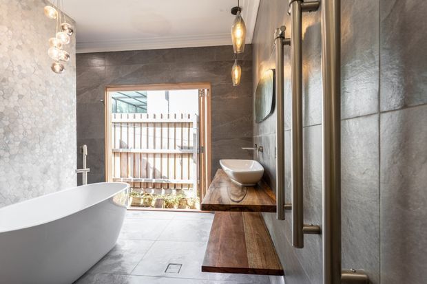 A contemporary bathroom featuring a freestanding white bathtub, a timber floating vanity with a vessel basin, dark large-format wall tiles, pale floor tiles, glass shower enclosure, and warm pendant lighting. A timber privacy fence and outdoor area are visible through the window.