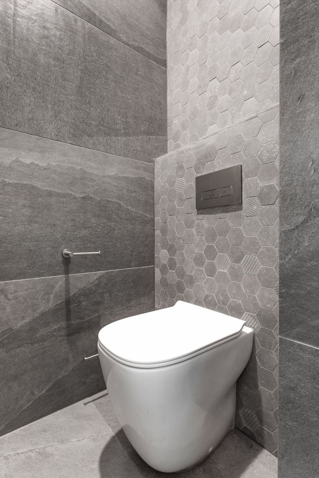 Close-up view of a contemporary bathroom toilet area. A wall-hung white toilet sits against large-format dark grey stone-look wall and floor tiles, with a vertical feature strip of small light grey hexagonal tiles and a dark flush plate. Minimal chrome toilet-roll holder and clean geometric detailing create a restrained monochrome finish.