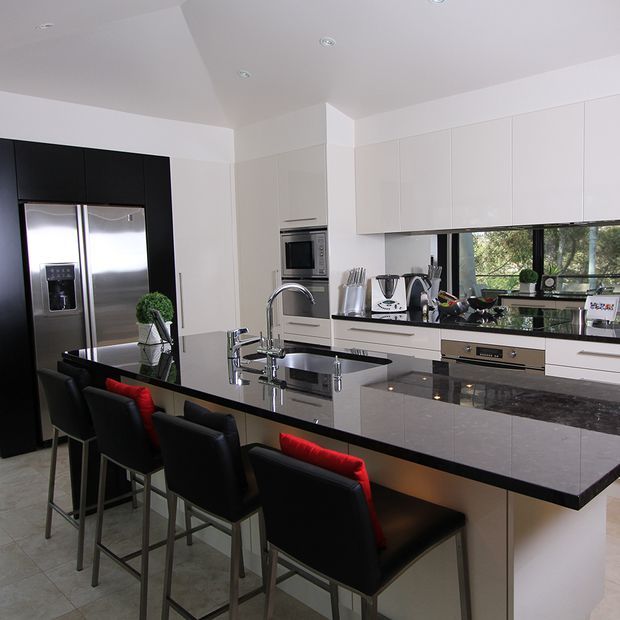 A contemporary kitchen with extensive white flat-panel cabinetry, a glossy black island and benchtops, integrated stainless-steel appliances, and a mirrored backsplash reflecting the garden outside. The large island includes a sink and seating for several people on black bar stools with red cushions. Recessed ceiling lights and broad glazing create a bright, polished space.