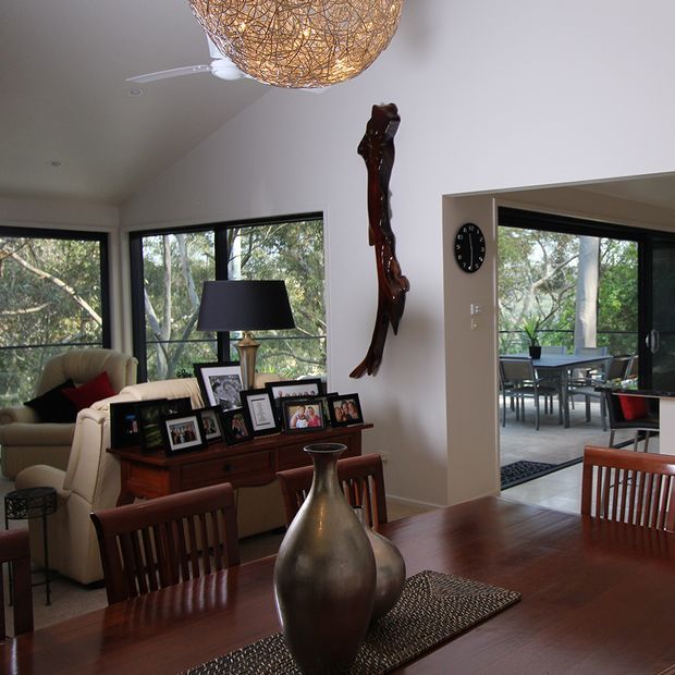 A warm, naturally lit open-plan dining and living area. The foreground features a polished dark timber dining table with wooden chairs, a large metallic decorative vase and woven runner. Beyond it is a living room with a cream sofa, dark timber sideboard, framed family photographs and table lamp. Floor-to-ceiling corner windows overlook trees, while a wide opening connects to a secondary dining or outdoor room. A sculptural pendant light and organic wall artwork add visual interest.