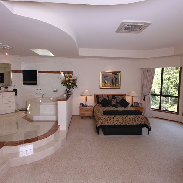 A spacious traditional bedroom with an integrated open ensuite area. The room features a large bed with patterned bedding, bedside tables and lamps, framed artwork, beige carpet, and a large curtained window. The ensuite includes a raised corner bathtub, tiled walls and floor, vanity cabinetry, and a curved architectural ceiling with recessed lighting.