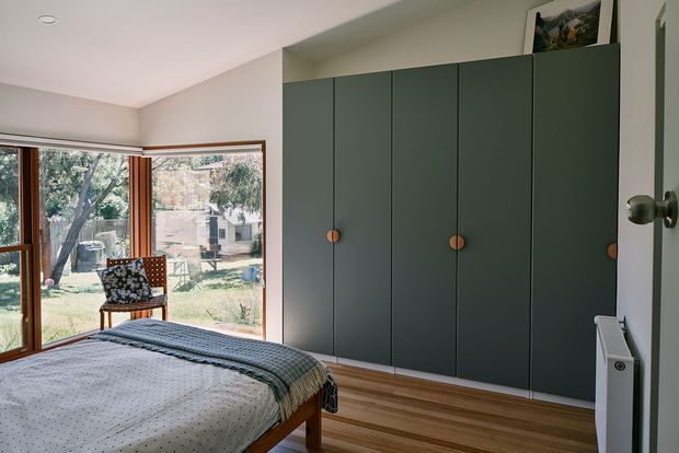 A calm, contemporary bedroom with a low timber bed, white dotted bedding, and a folded blue-grey throw. A large dark green built-in wardrobe with simple vertical doors and round timber knobs spans one wall. Timber-framed corner glazing opens the room to a leafy garden, while pale walls and timber flooring create a warm, light-filled interior.
