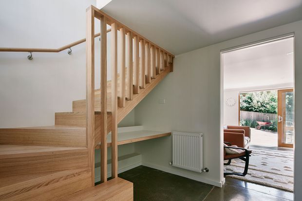 Interior view of a contemporary stairway and landing. A light timber staircase with open vertical balustrade rises beside a pale painted wall, with a timber handrail, built-in landing desk or shelf, white radiator, and dark flooring. An open doorway leads to a bright living area and outdoor patio beyond.