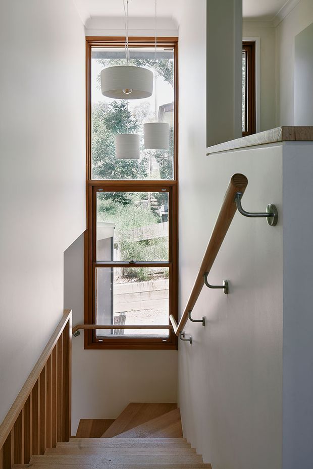 A bright, narrow interior stairway with warm timber treads, a timber balustrade, and a matching timber handrail mounted on a white wall with metal brackets. A tall timber-framed window floods the stairwell with natural light, while three pendant lights hang in front of the glazing. The space has simple white walls, restrained detailing, and a clean contemporary feel.