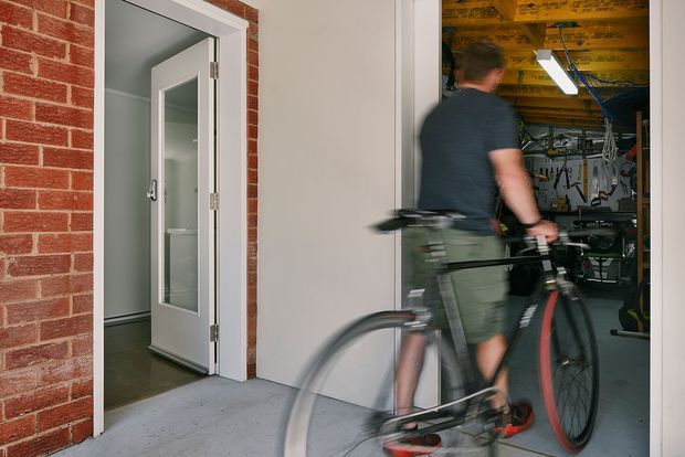 A photograph showing a cyclist wheeling a bicycle into an open garage or workshop. The scene includes exposed red brickwork, a white-framed glazed door leading to an interior room, and a garage interior with visible timber ceiling structure, fluorescent lighting, tools, and bicycle storage. Motion blur conveys movement into the garage.