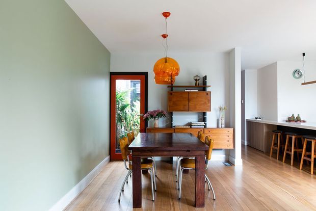 Bright open-plan mid-century-inspired dining and kitchen interior. A dark timber dining table and chairs sit in the foreground beneath a prominent amber pendant light, with a wall-mounted timber sideboard and shelving behind. The adjoining kitchen has a long island with dark countertop and timber cabinetry, while pale green and white walls, timber flooring, and a glazed exterior door create an airy, warm space.
