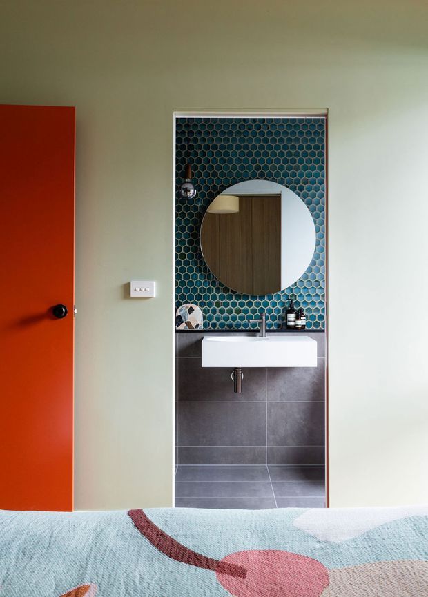 View from a bedroom toward an open bathroom vanity area. The compact bathroom features a white wall-mounted basin, dark grey floor and lower wall tiles, vivid blue-green hexagonal mosaic tiles on the upper wall, a round mirror, black tapware and accessories, and a bright orange-red door. The foreground shows the edge of a bed, confirming the bathroom opens directly from the bedroom.