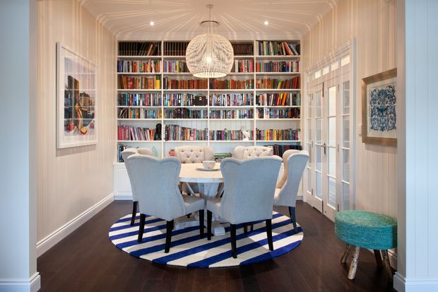 A refined, traditional dining room or library dining space with a round table surrounded by upholstered blue dining chairs, set on a bold blue-and-white striped circular rug. The back wall is fully fitted with floor-to-ceiling white bookshelves filled with books. White panelled walls, decorative ceiling moulding, French doors with glazed panes, framed artwork, and a sculptural pendant light create a bright, elegant interior. Dark timber flooring provides contrast, while a turquoise upholstered stool adds a small accent.