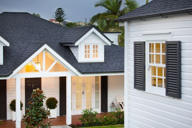 Exterior view of a light-colored, cottage-like residential building with overlapping gabled roof forms, dark shingle roofing, white horizontal weatherboard cladding, black louvred shutters, and multi-pane windows. A covered entry porch and landscaped planting are visible, with tropical palms in the background.
