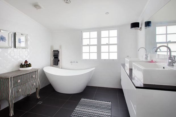 Bright, spacious contemporary bathroom with a freestanding white oval bathtub, twin white rectangular vessel basins on floating cabinetry, a large wall mirror, chrome tapware, white tiled walls, and dark charcoal floor tiles. Multi-pane windows provide abundant natural light, while a striped vanity cabinet and patterned bath mat add decorative contrast.