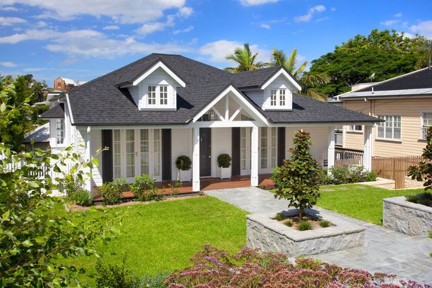 A bright exterior view of a detached cottage-style house with a dark shingle gabled roof, white timber facade, decorative gable trusses, dormer windows, and a covered front veranda. The landscaped front garden includes manicured lawn, raised stone planters, paved paths, and tropical vegetation.