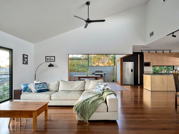 Open-plan contemporary living area with a large white sectional sofa, timber coffee table, hardwood flooring, high white ceiling, and black ceiling fan. The adjoining kitchen has dark cabinetry, light timber-toned joinery, stainless-steel appliances, and track lighting. Glazing and a wide opening connect the living space to a covered outdoor dining area and greenery beyond.