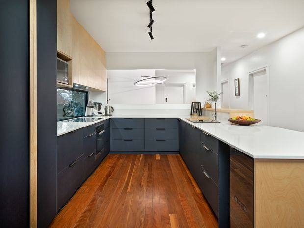 A bright contemporary U-shaped kitchen with dark navy-blue lower cabinetry, pale timber upper cabinetry and island panel, white solid-surface benchtops, and warm polished timber flooring. Integrated appliances, a window, pendant lighting, and recessed ceiling lights complete the clean, functional space.