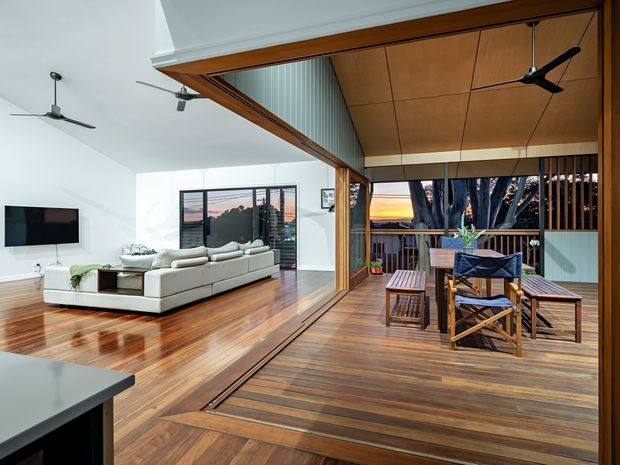 Bright open-plan living room opening onto a timber-lined covered deck through wide glazed doors. The interior features polished timber flooring, a white sectional sofa, wall-mounted television, ceiling fans, and large sliding glass panels. The deck has timber decking, a warm timber soffit, outdoor dining furniture, vertical privacy screening, and views toward a sunset and trees.