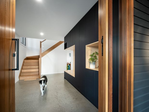 A contemporary residential entryway viewed through a timber-framed front door. The foyer features a polished concrete floor, a black built-in joinery wall with recessed timber display niches, a timber staircase with matching handrail, white walls and ceiling, and dark horizontal exterior cladding visible at the right edge. A dog is present in the hallway.