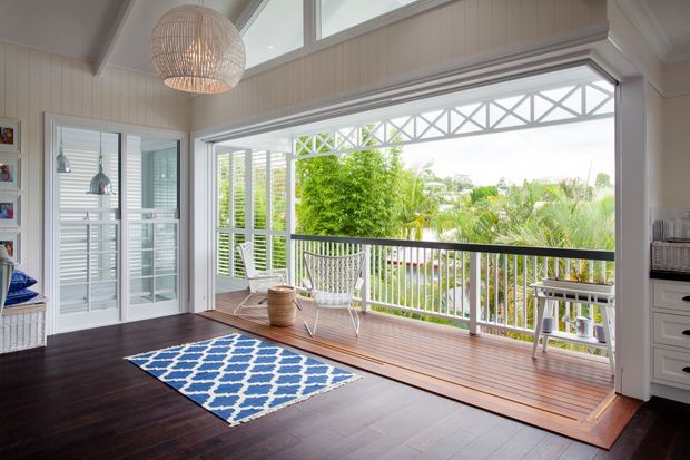 Bright coastal-style sunroom or living area opening onto a covered timber deck through large bifold or sliding glazing. The room has dark timber flooring, white vertical wall panelling, plantation shutters, a woven pendant light, and a blue-and-white patterned rug. The deck features white railings, outdoor seating, and lush tropical greenery beyond.