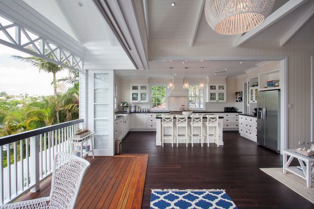 Bright open-plan kitchen and dining area opening onto a covered timber balcony or terrace. The space features white shaker-style cabinetry, a large island with white cross-back stools, dark timber flooring, light grey vertical wall panelling, exposed white ceiling beams, stainless-steel appliances, and woven pendant lighting. Tropical planting is visible beyond the balcony railing.