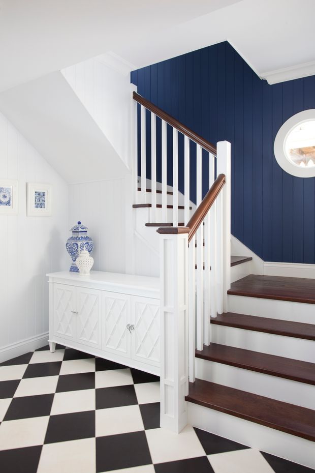 Bright, refined stairway and landing interior with dark stained timber stair treads and handrails, white painted balustrades and panelling, a navy blue vertical-panel feature wall, and a bold black-and-white checkerboard floor. A white decorative console with geometric door fronts sits beneath framed artwork, while a round window adds architectural interest.