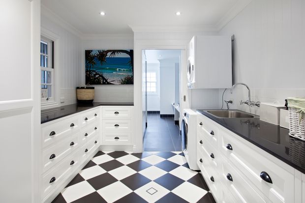 Bright, narrow traditional-style laundry room with extensive white built-in cabinetry, black stone-look benchtops, a stainless-steel sink, washing appliances, and a bold black-and-white checkerboard tile floor. White wall panelling, decorative trim, sash windows, and coastal artwork create a crisp, classic character. A hallway or utility space is visible beyond the doorway.