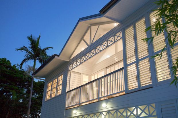 Twilight exterior view of a raised, light-coloured weatherboard house with a broad covered upper-level verandah or balcony. The facade features decorative geometric balustrade panels, louvred shutters, white-painted timber detailing, a pitched roof with deep eaves, and warm integrated lighting. Tropical planting and a palm tree frame the home.