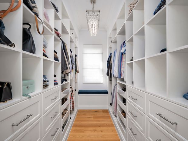 A bright, narrow walk-in wardrobe with floor-to-ceiling white built-in shelving, hanging rails, open cubbies, and numerous drawers on both sides. Clothing, shoes, and handbags are neatly arranged throughout the storage units. A timber floor leads to a window seat with a dark blue cushion beneath a white window, while a crystal-style pendant light provides a decorative focal point.
