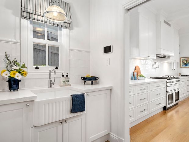 Bright, traditional-style utility/laundry area with a deep white farmhouse sink, white paneled cabinetry, tiled walls, timber flooring, and a window with striped Roman blind. The space opens directly into a white kitchen with extensive cabinetry, a freestanding range cooker, and stainless-steel appliances. Polished chrome tapware and dark blue accents add contrast.