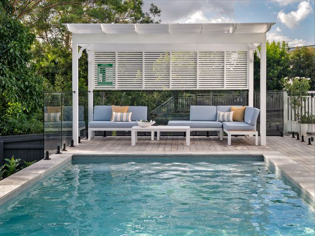 A landscaped outdoor pool area with a raised timber deck, glass balustrades, and a white pergola sheltering a built-in outdoor lounge. The seating includes a sofa, armchairs, cushions, and a low coffee table overlooking the swimming pool. White horizontal louvres provide privacy and shade, with dense greenery surrounding the space.