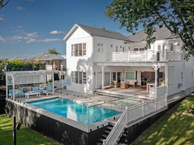 Exterior view of a large white weatherboard multi-level home with a gabled tiled roof, wraparound balcony, covered outdoor lounge and a raised swimming pool with glass and white balustrades. The house has traditional detailing and extensive indoor-outdoor living areas, set in a leafy suburban environment.