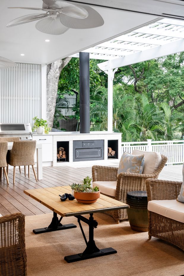 Bright, airy covered outdoor entertaining area with a tropical garden outlook. The space combines a compact outdoor kitchen and built-in barbecue/fireplace surround with a dining counter, wicker lounge seating, a timber coffee table, white vertical wall cladding, and a white pergola-style roof structure. Timber decking, woven furnishings, and lush planting create a relaxed resort-like atmosphere.