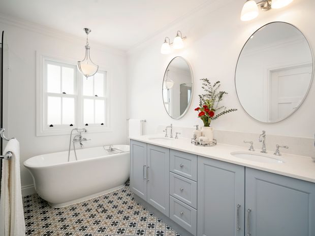Bright, spacious traditional bathroom with a freestanding white bathtub, double vanity, pale blue-grey cabinetry, white countertop, twin oval mirrors, classic chrome fixtures, and decorative patterned floor tiles. White walls, detailed trim, and pendant and vanity lighting create a refined, airy aesthetic.