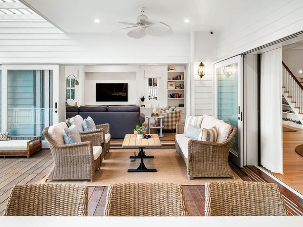Bright, airy coastal-style living room opening onto a glazed outdoor area. The room features white horizontal wall panelling, timber flooring, extensive sliding glass doors, woven rattan lounge furniture, a dark sofa, built-in shelving, a wall-mounted television, and a ceiling fan. A stairway and adjoining space are visible through the open doors on the right.