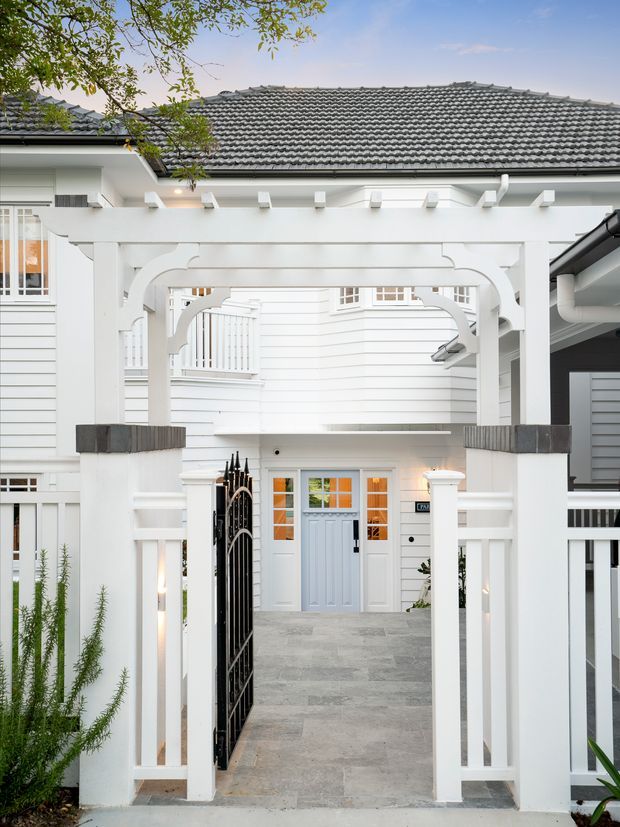 A front entry courtyard of a white weatherboard residence, framed by a substantial white timber pergola and arched gate structure. The entry path is paved in large grey stone slabs and leads to a pale blue front door with glazed sidelights and warm interior light. A black metal pedestrian gate, white fencing, decorative timber brackets, and a grey tiled pitched roof are visible, with limited greenery around the edges.
