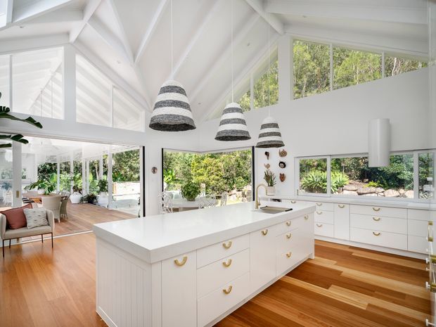 Bright open-plan white kitchen with a large central island, extensive white cabinetry, timber flooring, and generous glazing overlooking lush greenery. Exposed white structural beams and a high pitched ceiling create an airy, light-filled interior. Three woven pendant lights hang above the island, while a living area is visible beyond the kitchen.