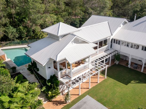 Aerial view of a large white weatherboard house set in a leafy, wooded site. The home has multiple pitched and gabled corrugated metal roof sections, extensive verandahs with white balustrades, a covered pergola, and a central grass courtyard. A rectangular swimming pool and paved terrace are visible beside the house.