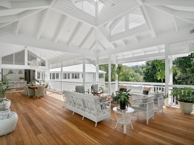 A bright, spacious enclosed veranda or sunroom with a white-painted timber structure, vaulted roof, exposed rafters, clerestory-style louvred openings, and a central skylight. The room features warm timber decking, white wicker-style lounge furniture, a dining setting, extensive glazing, and abundant indoor plants, with leafy greenery visible outside. The overall atmosphere is airy, relaxed, and coastal-inspired.